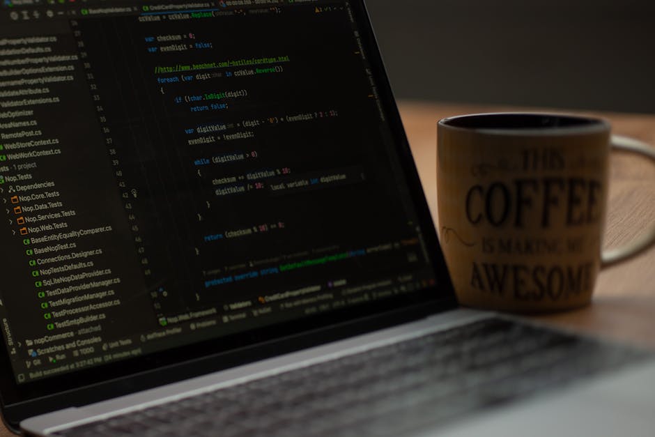 Close-up of an open laptop displaying lines of code on a dark background, situated on a wooden desk beside a ceramic coffee mug with black text that reads 'This coffee is maker me awesome.' In the bac